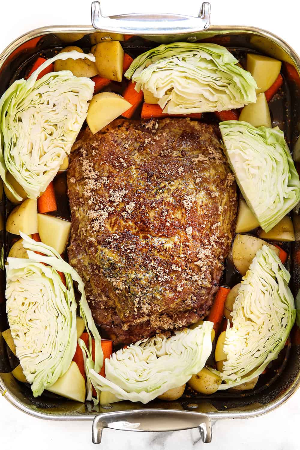showing how to make oven corned beef by adding corned beef brisket to a roasting pan and surrounding by carrots, potatoes and sliced cabbage