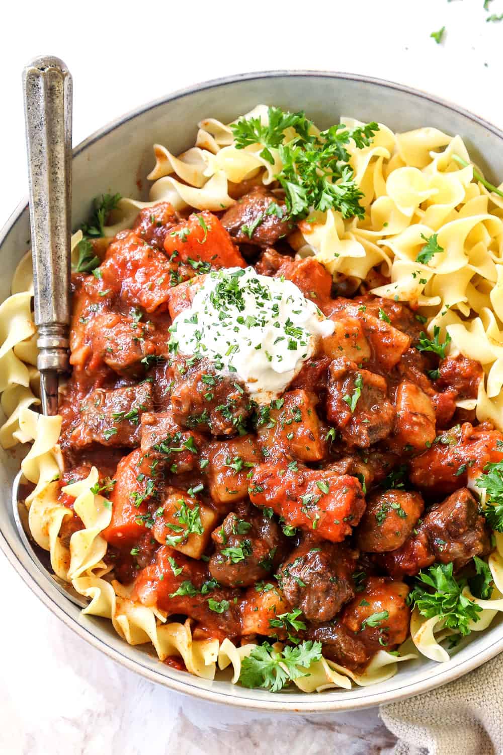 showing how to serve Hungarian Goulash recipe by adding sour cream to goulash in a bowl of egg noodles