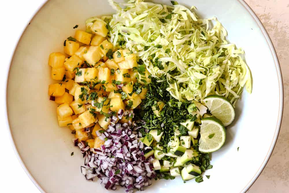 showing how to make shrimp taco recipe by making slaw by adding pineapple, cabbage, cucumbers, red onions and cilantro to a white bowl