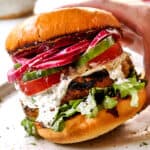 showing toppings for lamb burger Greek style by topping buns with arugula, lamb patties, tomatoes, cucumbers, tzatziki and red onions