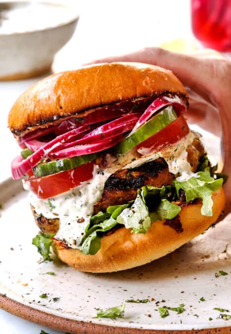 showing toppings for lamb burger Greek style by topping buns with arugula, lamb patties, tomatoes, cucumbers, tzatziki and red onions