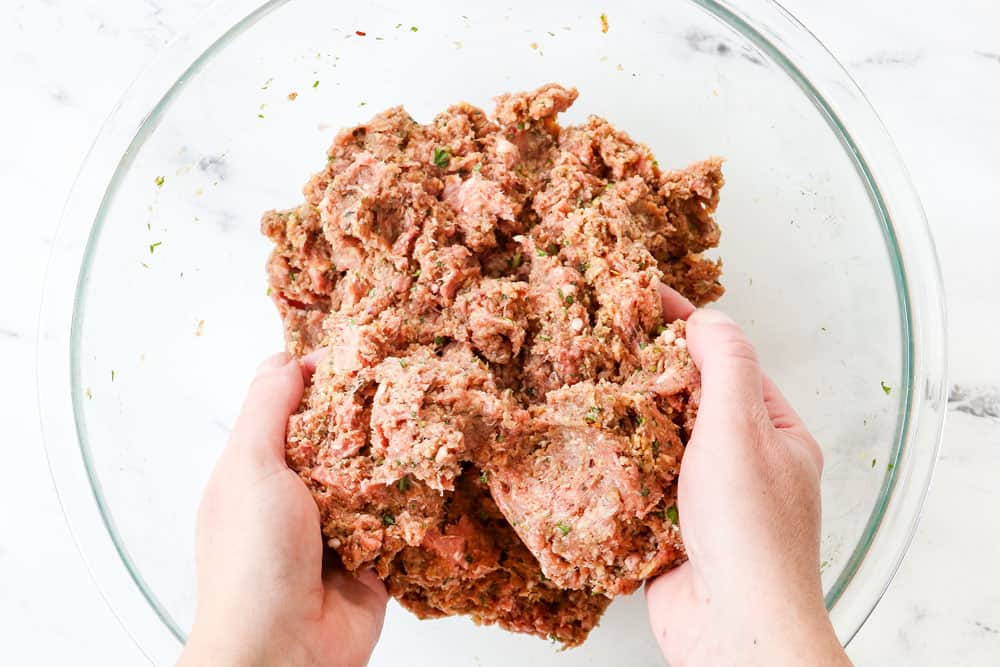 showing how to make Greek Meatball recipe (Keftedes) by mixing  ground lamb, ground beef, eggs, breadcrumbs, lemon juice, garlic, onion, basil, dill, oregano, mint to a glass bowl with two hands