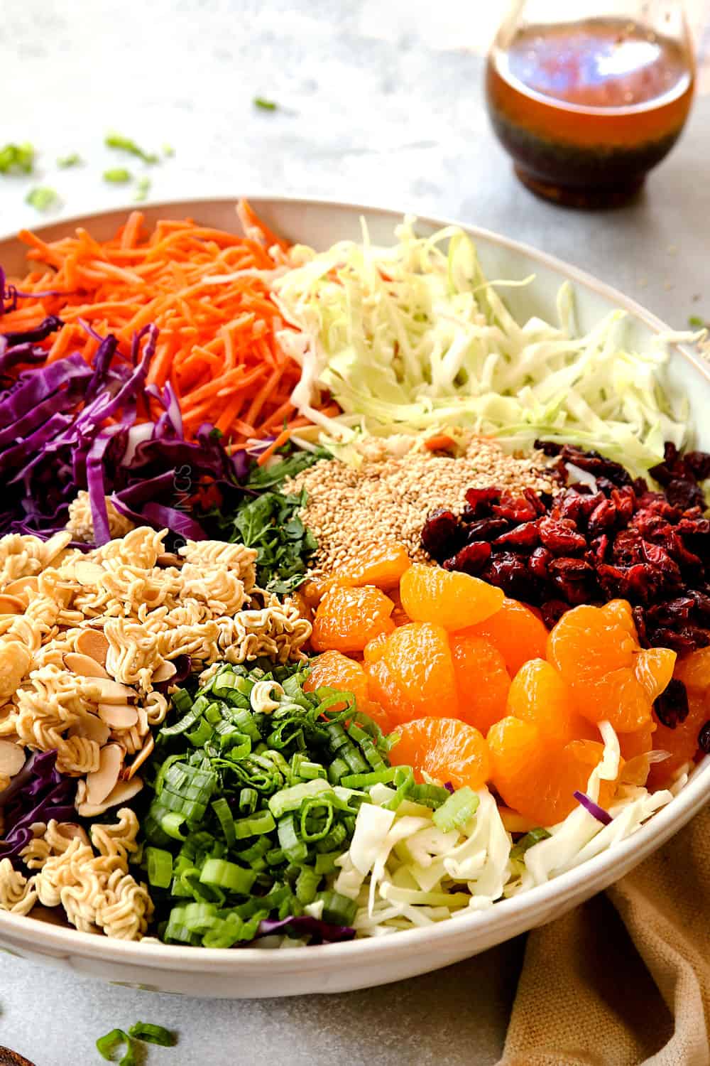 showing how to make Ramen Noodle Salad recipe with side view of ingredients in a bowl: cabbage, carrots, ramen noodles, almonds and optional toppings of sunflower seeds, Mandarin oranges, green onions and dried cranberries