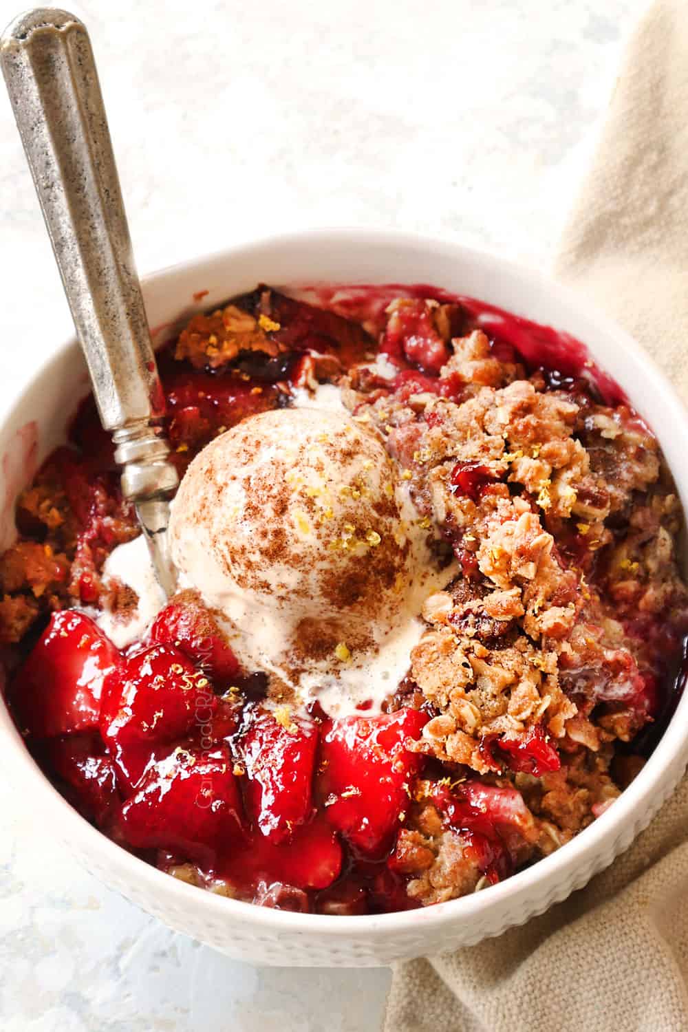 showing how to serve strawberry crisp (crumble) by adding to a white bowl topped with vanilla ice cream and cinnamon