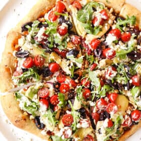 showing how to make Greek pizza recipe by adding arugula, tomatoes, cucumbers and tzatziki to the top of the baked pizza