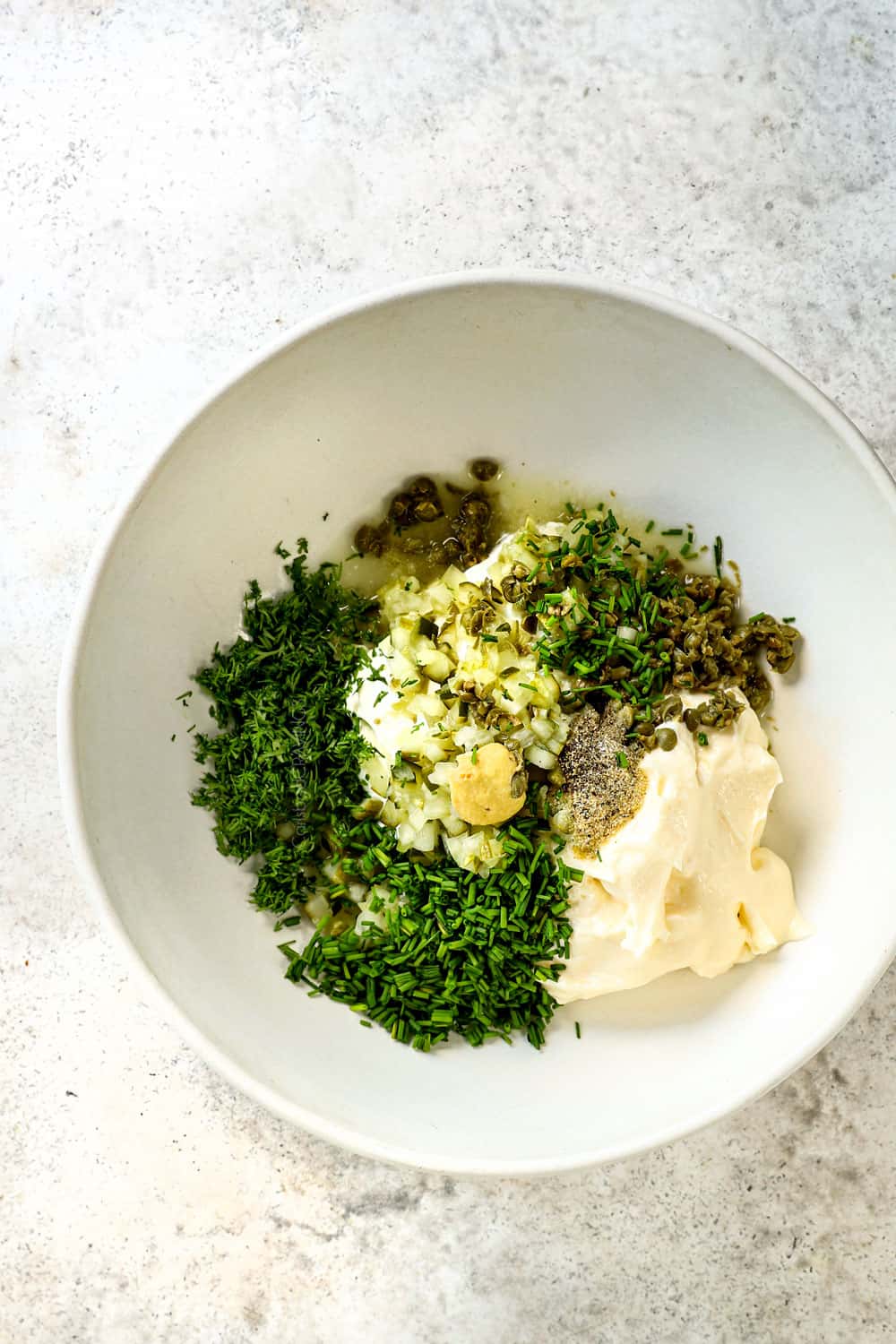 showing ingredients in tartar sauce by adding mayonnaise, diced pickle, diced capers, chopped dill, and lemon juice to a white bowl