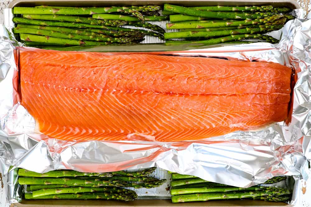 showing how to make baked lemon pepper salmon recipe by adding salmon to a sheet pan with asparagus to bake in the oven