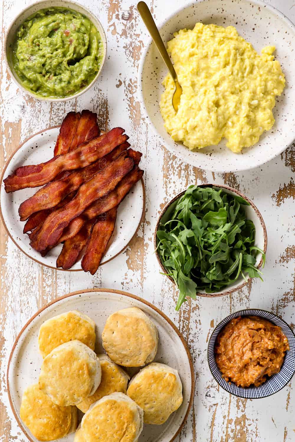 showing how to make breakfast sandwich recipe by showing ingredients: scrambled eggs, bacon, muffins, guacamole and arugula