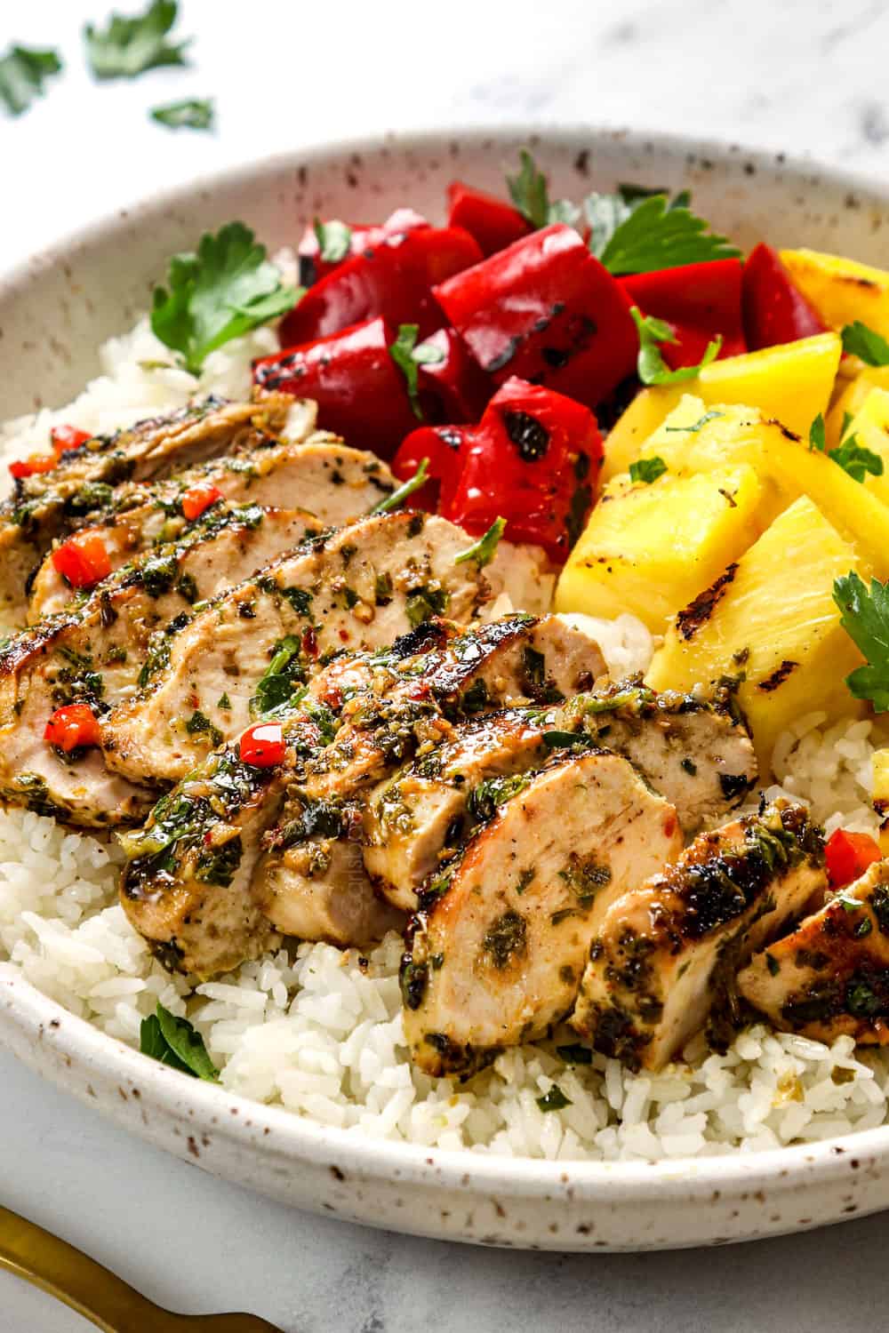 showing how to serve chicken chimichurri by adding it to a bowl with rice, pineapple and grilled bell peppers