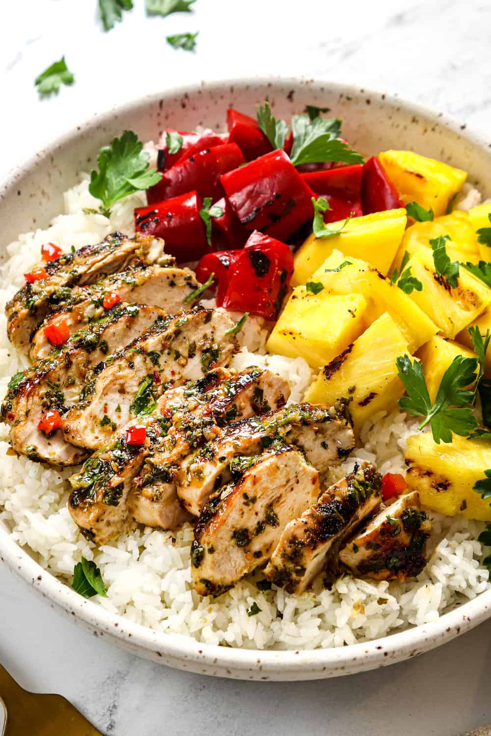 showing how to serve chicken chimichurri by adding it to a bowl with rice, pineapple and grilled bell peppers