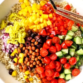 top view showing how to make couscous salad recipe by adding pearl couscous, tomatoes, cucumbers, chickpeas, bell peppers, red onions and olives to a bowl and tossing to combine