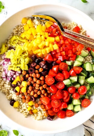 top view showing how to make couscous salad recipe by adding pearl couscous, tomatoes, cucumbers, chickpeas, bell peppers, red onions and olives to a bowl and tossing to combine