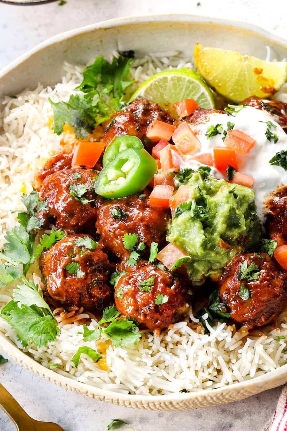 showing how to serve baked enchilada meatballs by adding to a bowl with rice and topping with sour cream, guacamole, tomatoes and cilantro