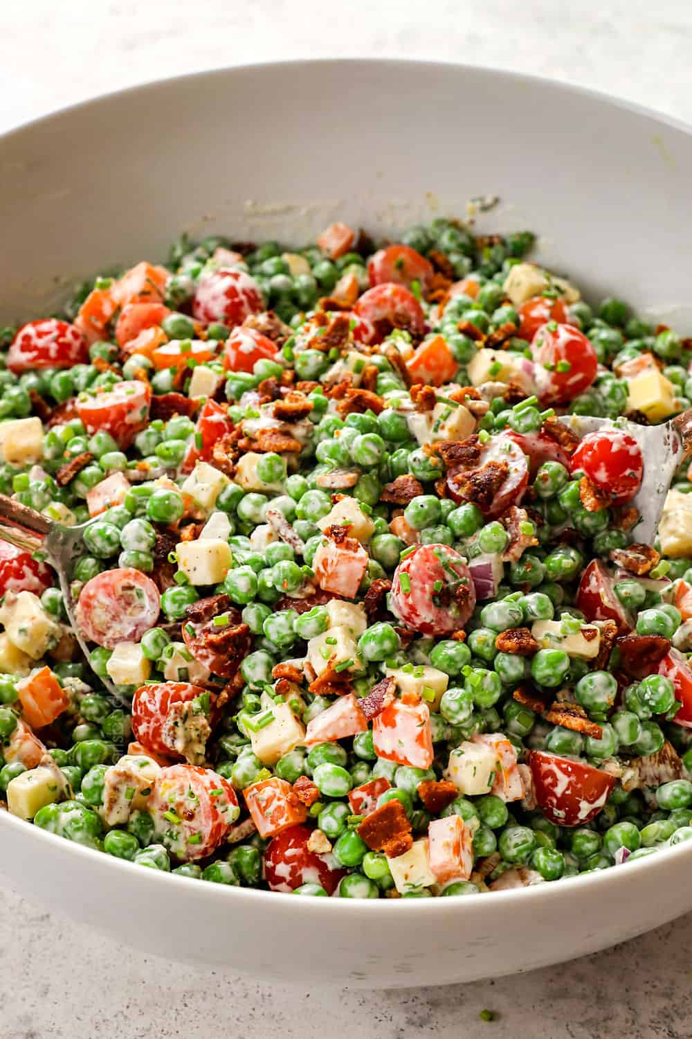 tossing pea salad together in a white bowl with green peas, bacon, tomatoes, red onions and bell peppers