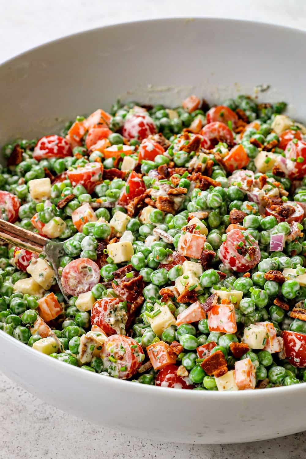 tossing pea salad together in a white bowl with green peas, bacon, tomatoes, red onions and bell peppers