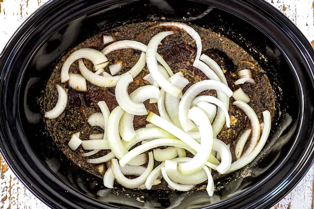 showing how to make Crockpot Philly Cheesesteak by adding chuck roast, braising liquid, seasonings and onions to a slow cooker
