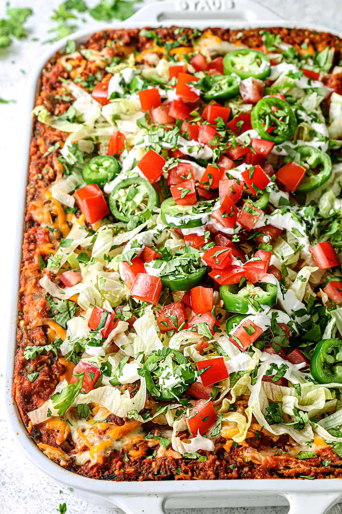 showing how to make taco lasagna by topping with lettuce, tomatoes, cilantro and jalapenos