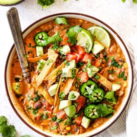 top view showing how to serve chicken tortilla soup by adding to a bowl and topping with tortilla strips, avocado, cilantro, jalapeno, fresh limes and tomatoes