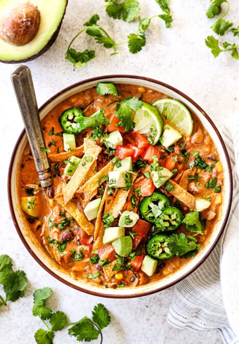 top view showing how to serve chicken tortilla soup by adding to a bowl and topping with tortilla strips, avocado, cilantro, jalapeno, fresh limes and tomatoes