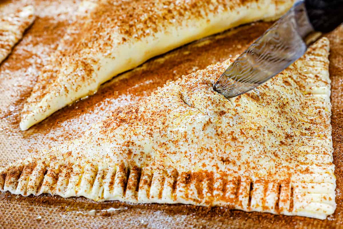showing how to make apple turnover recipe by dusting puff pastry with cinnamon and sugar, then making small slits in the top of the pastries with a pairing knife