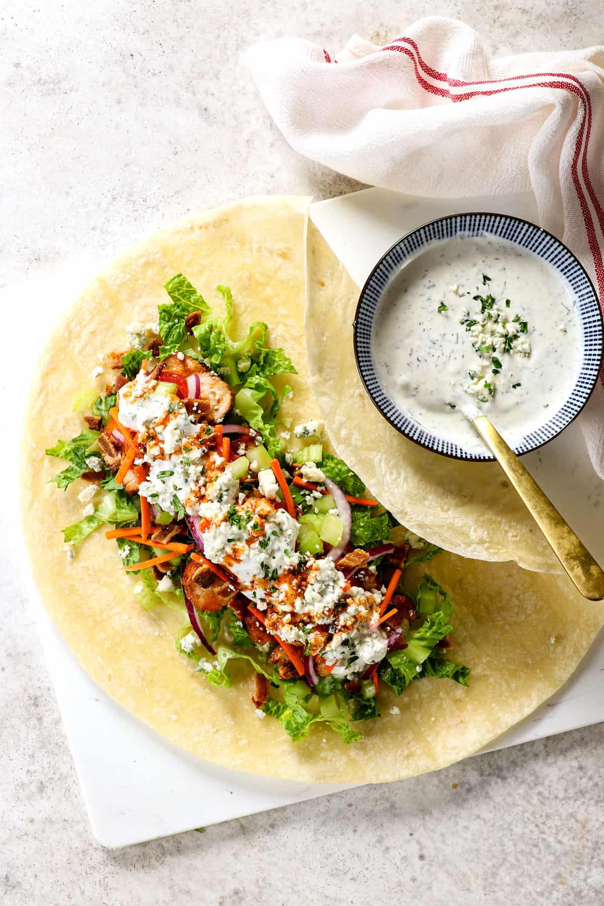 showing how to make buffalo chicken wraps by adding lining tortillas with lettuce, buffalo chicken, tomatoes, celery, carrots, red onions, bacon and blue cheese dressing