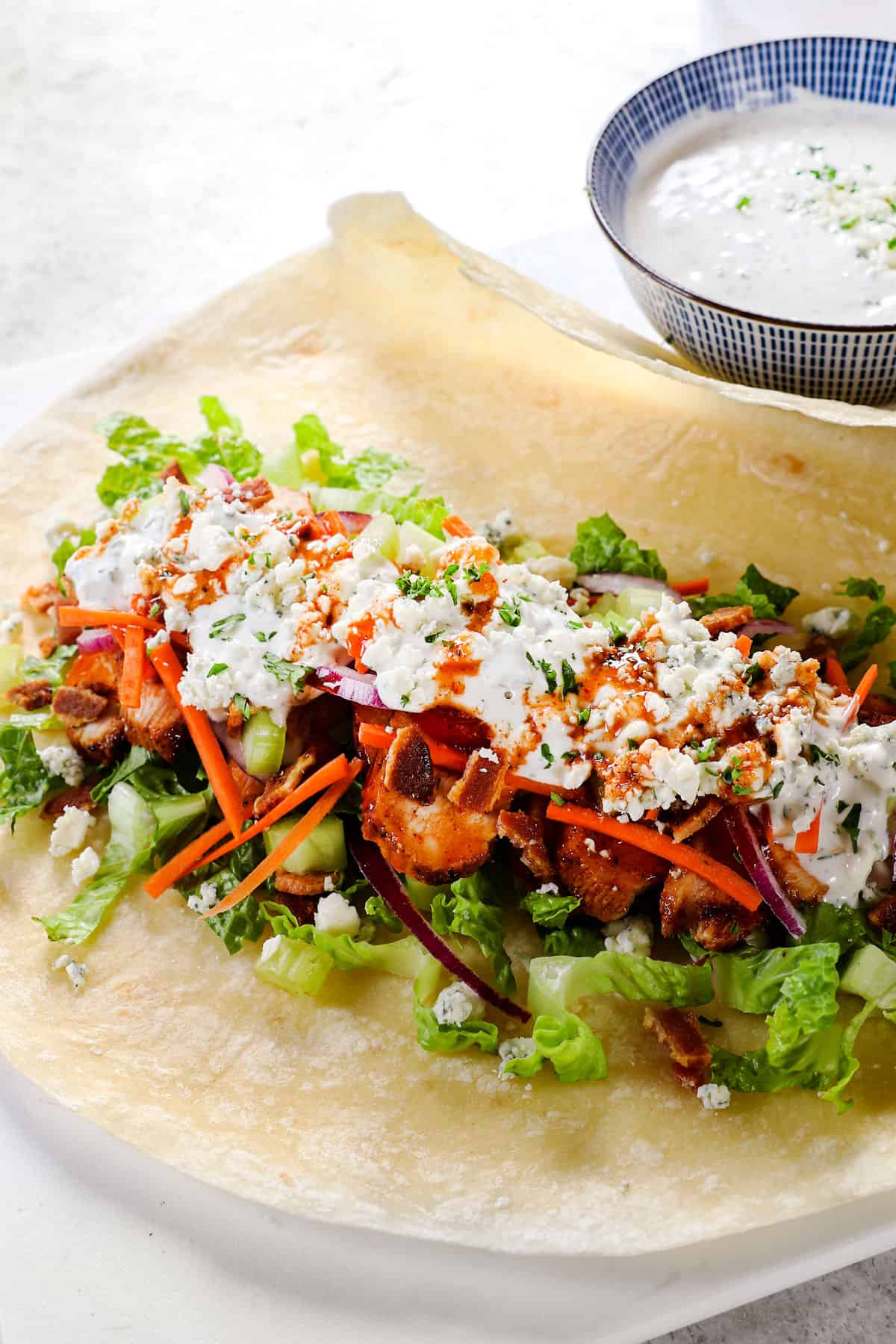 showing how to make buffalo chicken wraps by layering a tortilla with lettuce, buffalo chicken, red onions, carrots, celery and blue cheese dressing