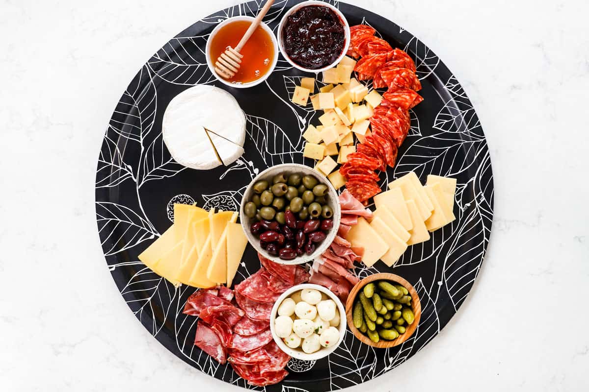 showing how to make a charcuterie board by adding jam, honey, mustard, olives and mini pickles on the board
