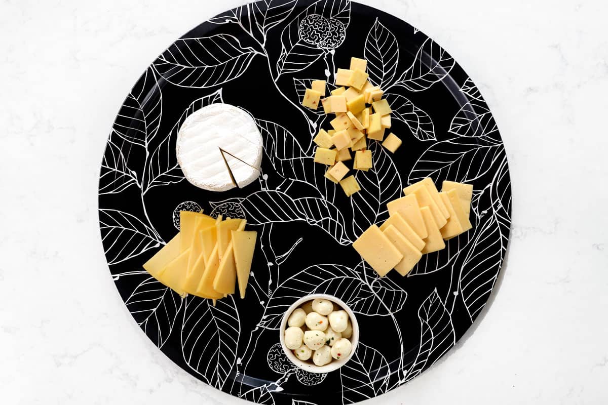 showing how to make a charcuterie board by adding soft cheese (brie), spreadable cheese (goat cheese), semi-soft cheese (Havarti) and hard cheese (Manchego) to a board
