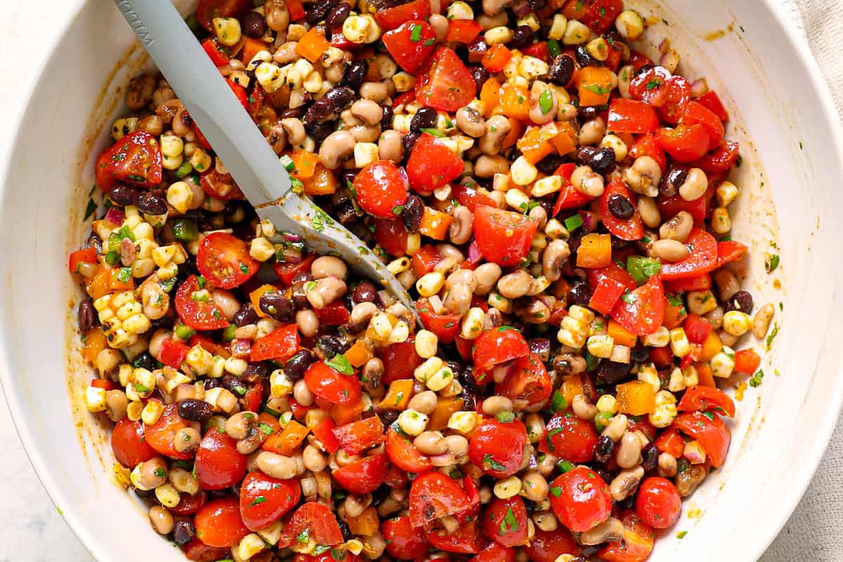 showing how to make easy cowboy caviar (Texas Caviar), by stirring black eyed peas, beans, tomatoes, corn, bell peppers, red onion and cilantro together in a bowl