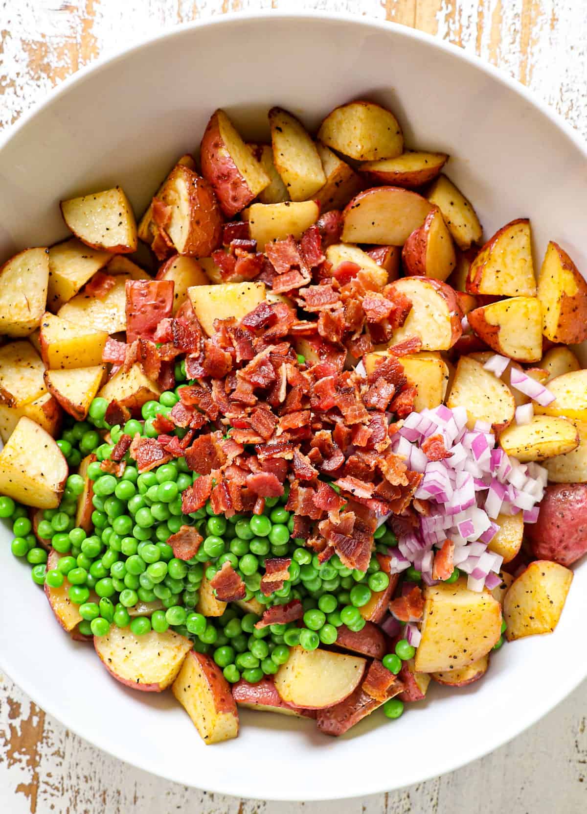 showing how to make red potato salad by adding red potatoes, peas, bacon and red onion to a large bowl