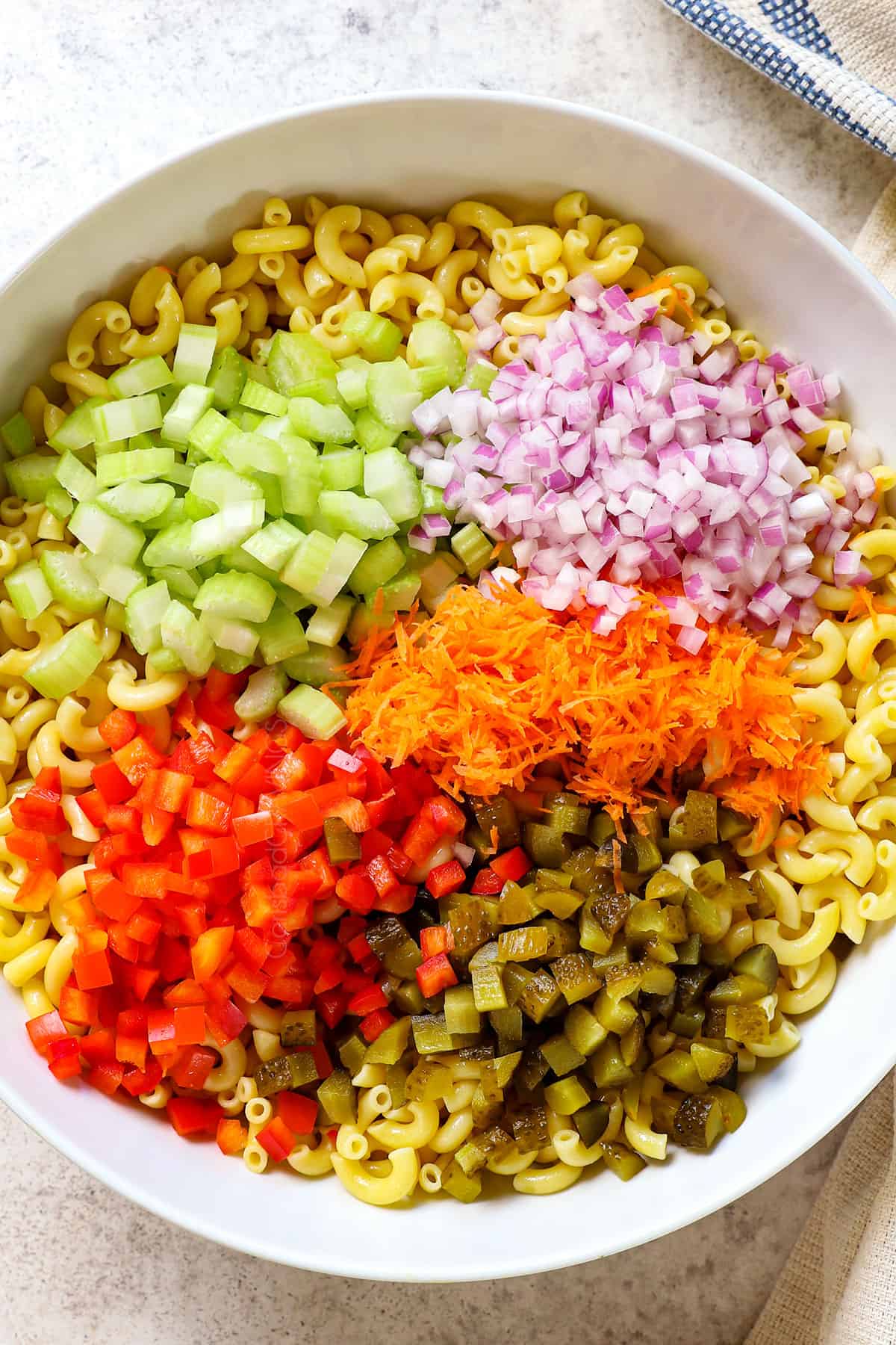 top view showing ingredients of macaroni salad recipe in a bowl:  macaroni, celery, red onions, pickles, shredded carrots