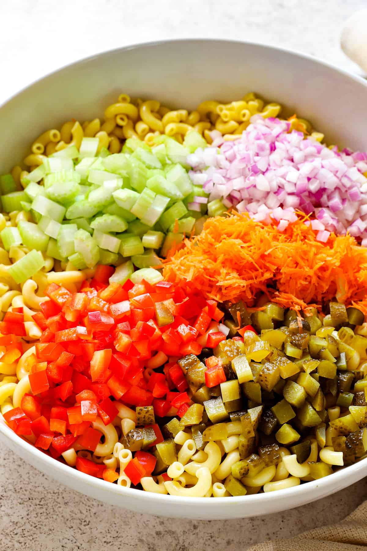 showing how to make Macaroni Salad by adding celery, bell peppers, pickles, shredded carrots and red onions to a bowl