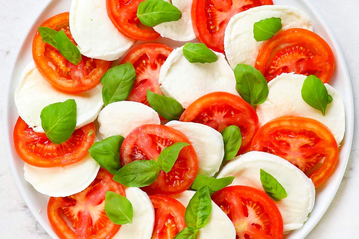 showing how to make Caprese Salad by layering mozzarella and tomatoes in an alternating row, then adding basil leaves