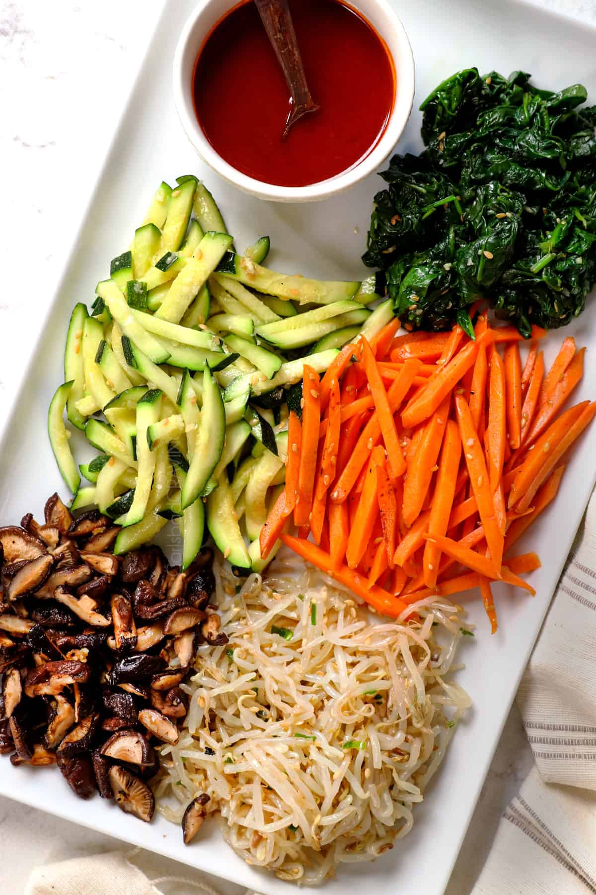 showing bibimbap recipe ingredients on a platter: mushrooms, bean sprouts, carrots, zucchini, spinach and bibimbap sauce