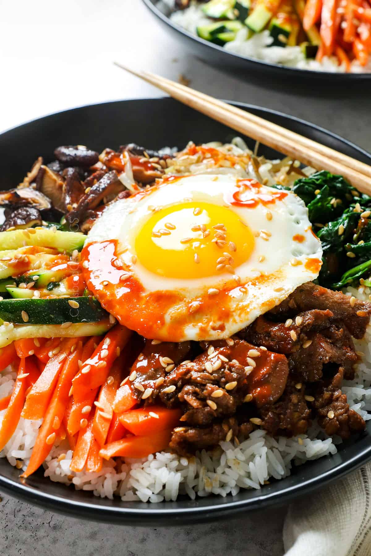 showing how to assemble bibimbap by adding carrots, zucchini, mushrooms, spinach, beef and egg to a bowl of rice