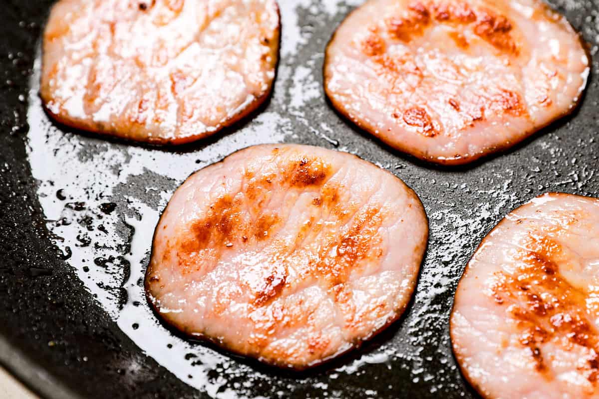 showing how to make Eggs Benedict recipe by cooking Canadian bacon in a skillet until crispy 