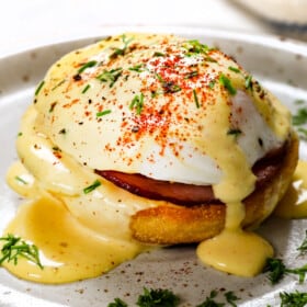 showing how to make Eggs Benedict recipe by assembling with English Muffin, Canadian Bacon, poached egg and hollandaise sauce