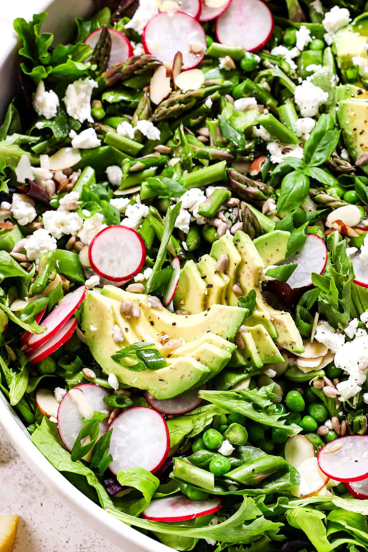 showing how to make spring salad by adding spring salad mix, asparagus, snap peas, radishes, avocado, goat cheese, almonds and sunflower seeds