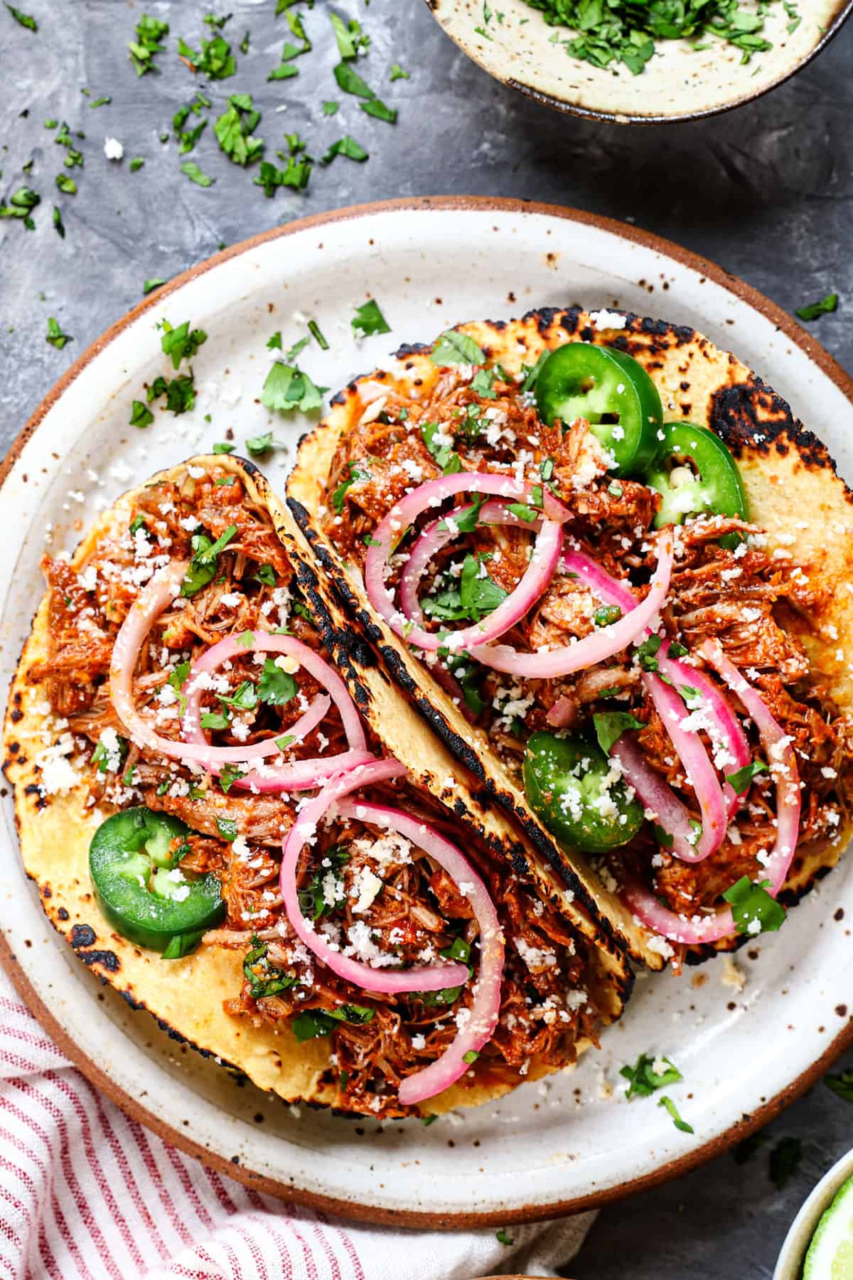 showing how to serve cochinita pibil by adding to warm corn tortillas and topping with pickled red onions, cilantro, and Cotija cheese