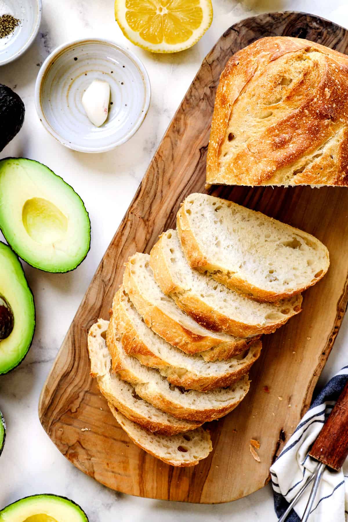 top view showing the ingredients for avocado toast:  sourdough bread on a cutting board, avocados, lemon juice, garlic, salt and pepper