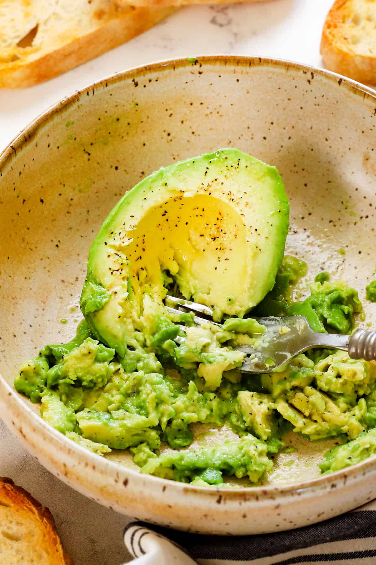 showing how to make avocado toast by mashing the avocado in a bowl with lemon juice, salt and pepper