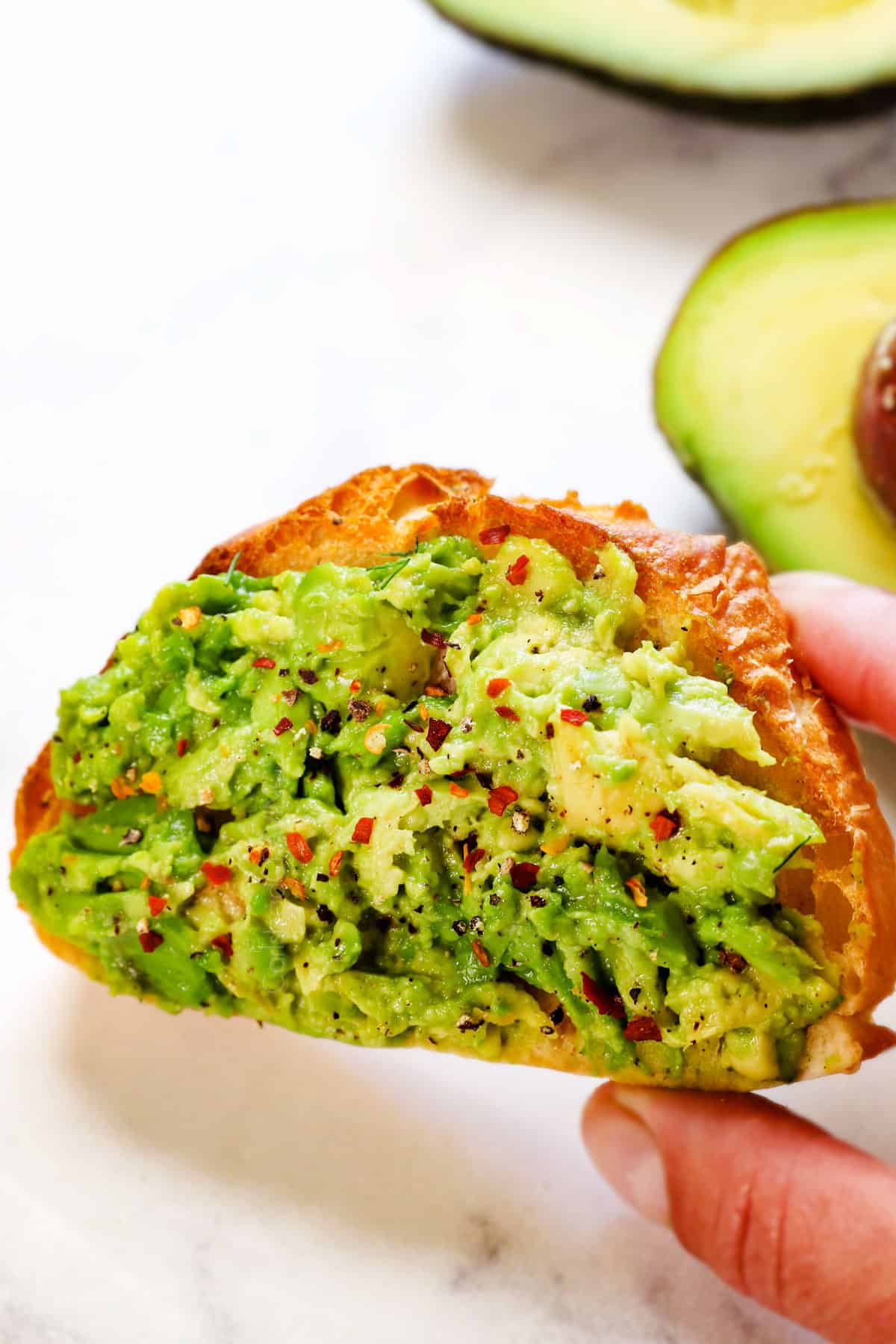 showing how to make avocado toast by spreading the mashed avocado onto the toast, then sprinkling with red pepper flakes