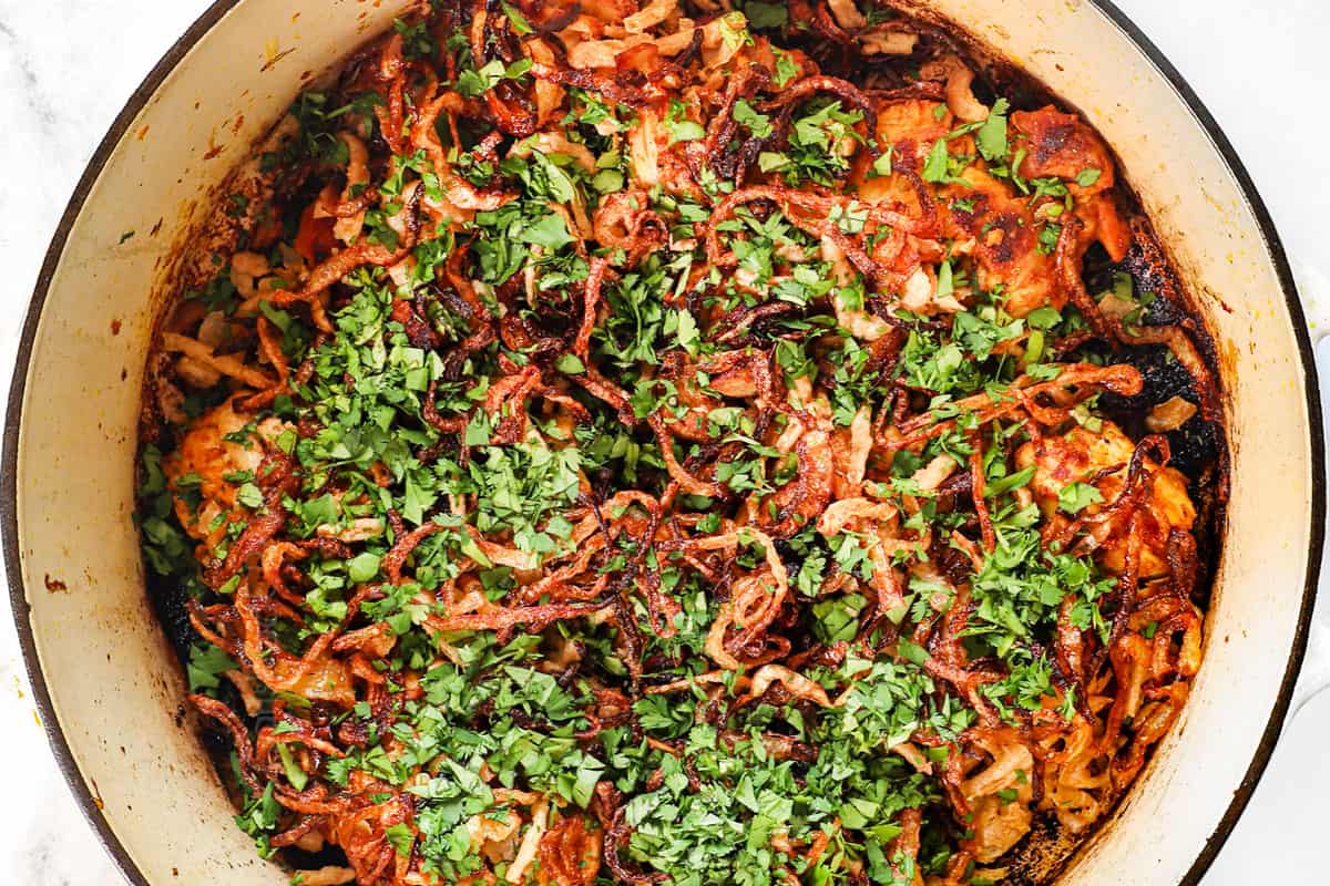 showing how to make biryani by layering the fried onions over the chicken, followed by the cilantro and mint