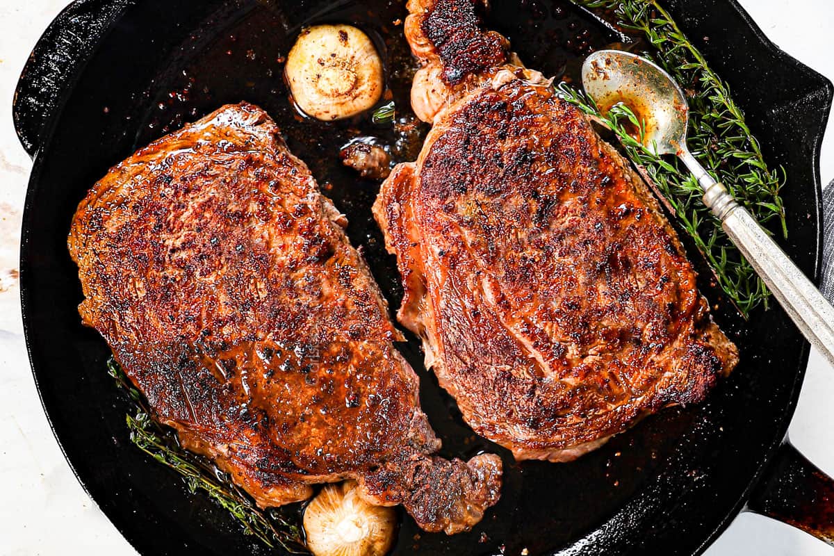 showing how to cook ribeye steak by adding garlic, rosemary and thyme to the cast iron skillet for pan searing