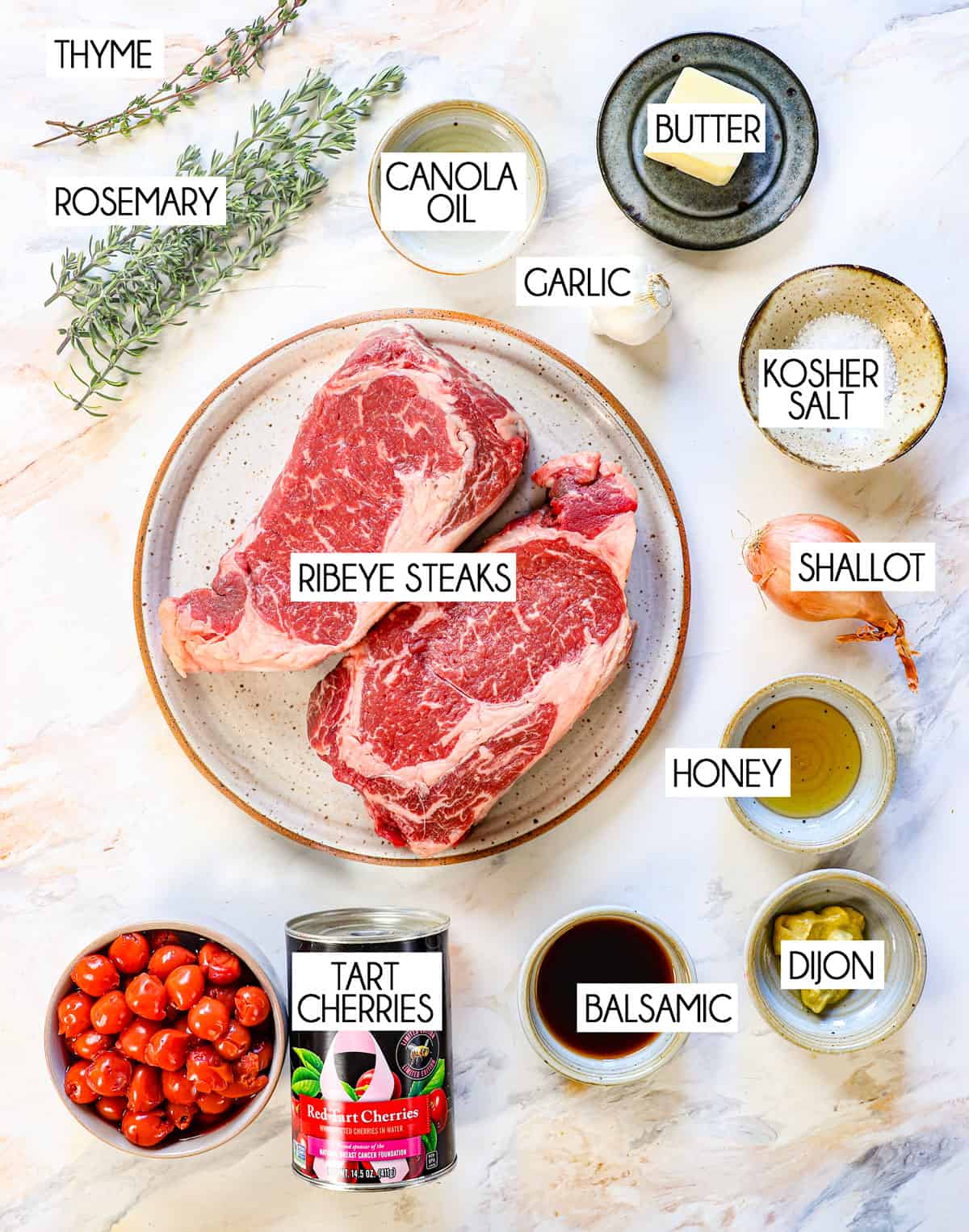 top view showing ingredients for ribeye steak recipe: two ribeye steaks, kosher salt, pepper, rosemary, thyme, garlic, canola oil, and butter