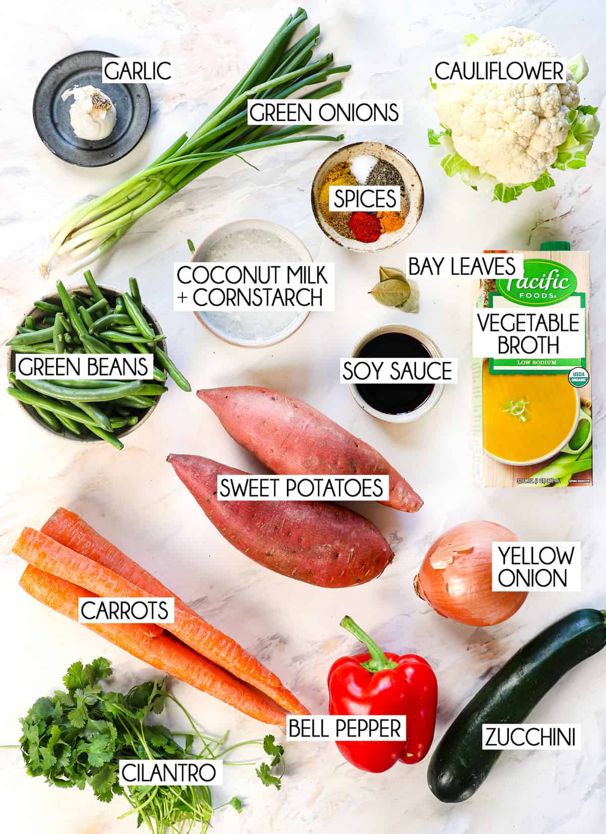 top view of vegetable soup ingredients: sweet potatoes, bell pepper, carrots, zucchini, onion, garlic, green beans, cauliflower, vegetable broth, coconut milk, spices