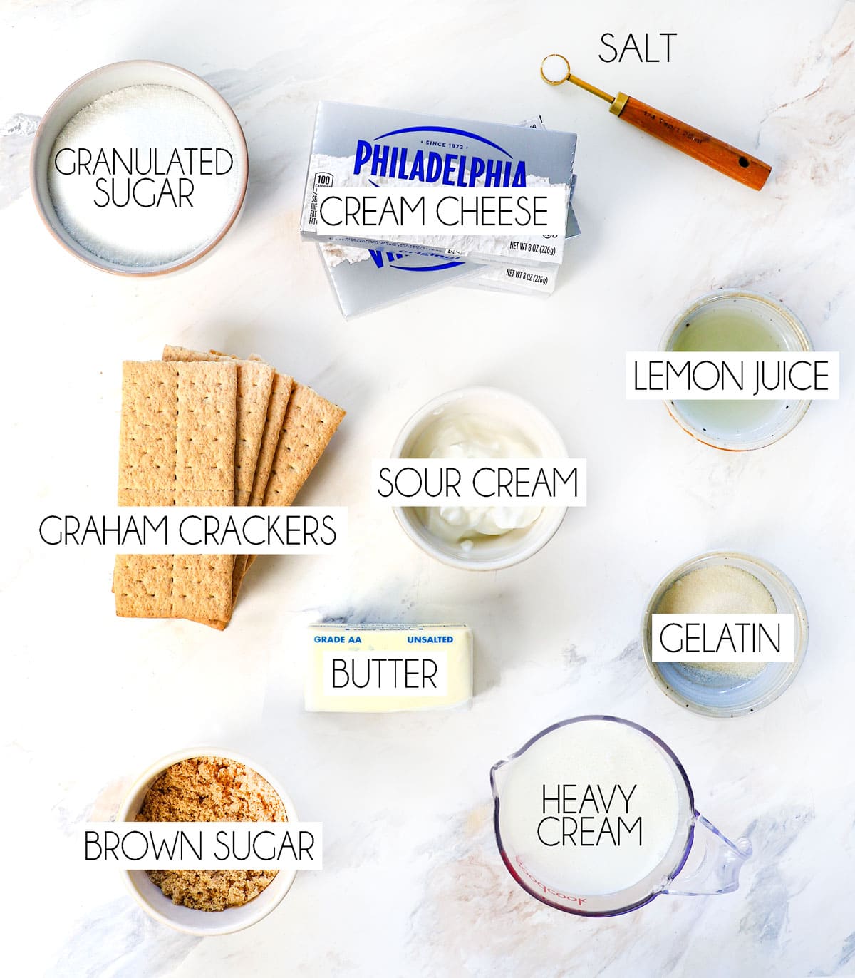 top view of no bake cheesecake ingredients: cream cheese, heavy cream, sugar, gelatin, lemon juice, sour cream, graham crackers, brown sugar, butter, and salt