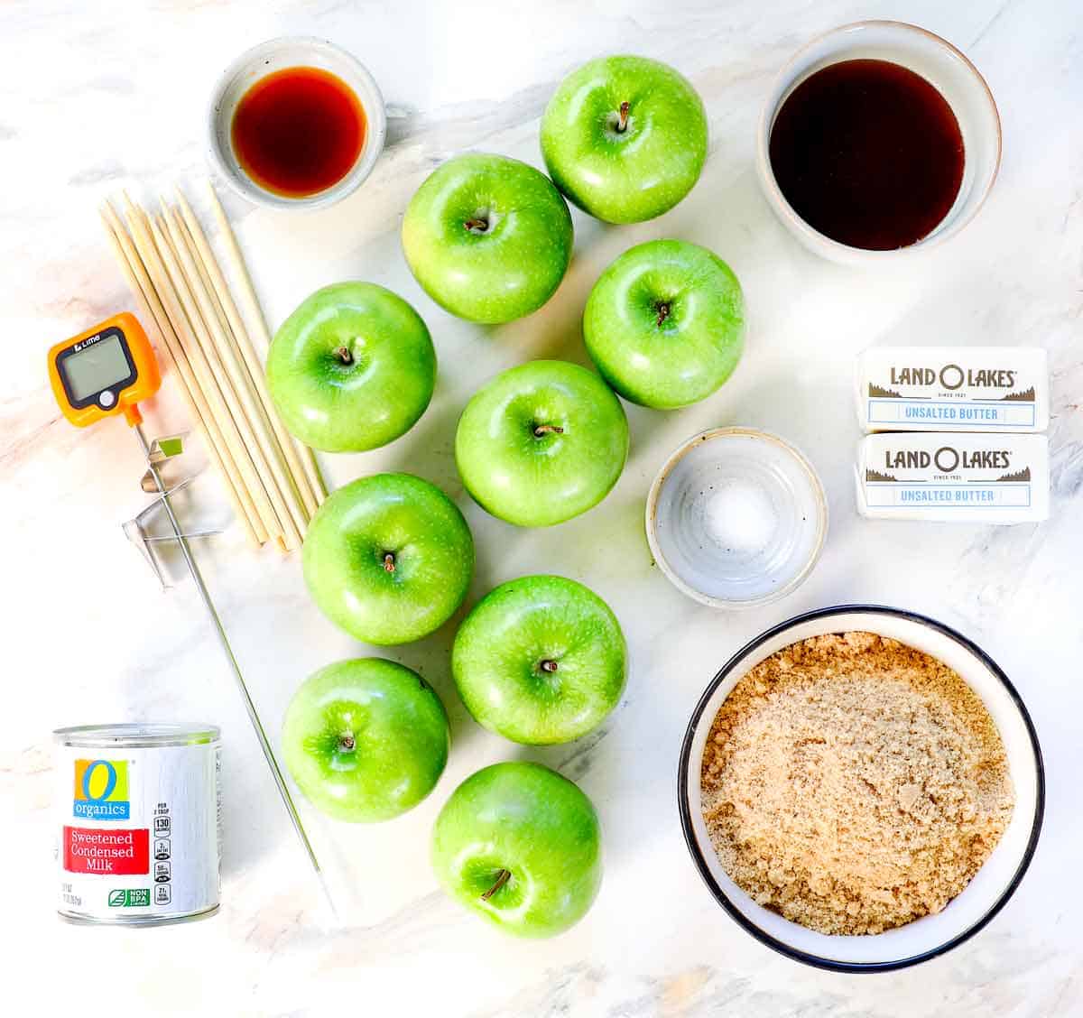 top view of caramel apple recipe ingredients: Granny smith apples, brown sugar, sweetened condensed milk, butter, vanilla, apple sticks, and candy thermometer