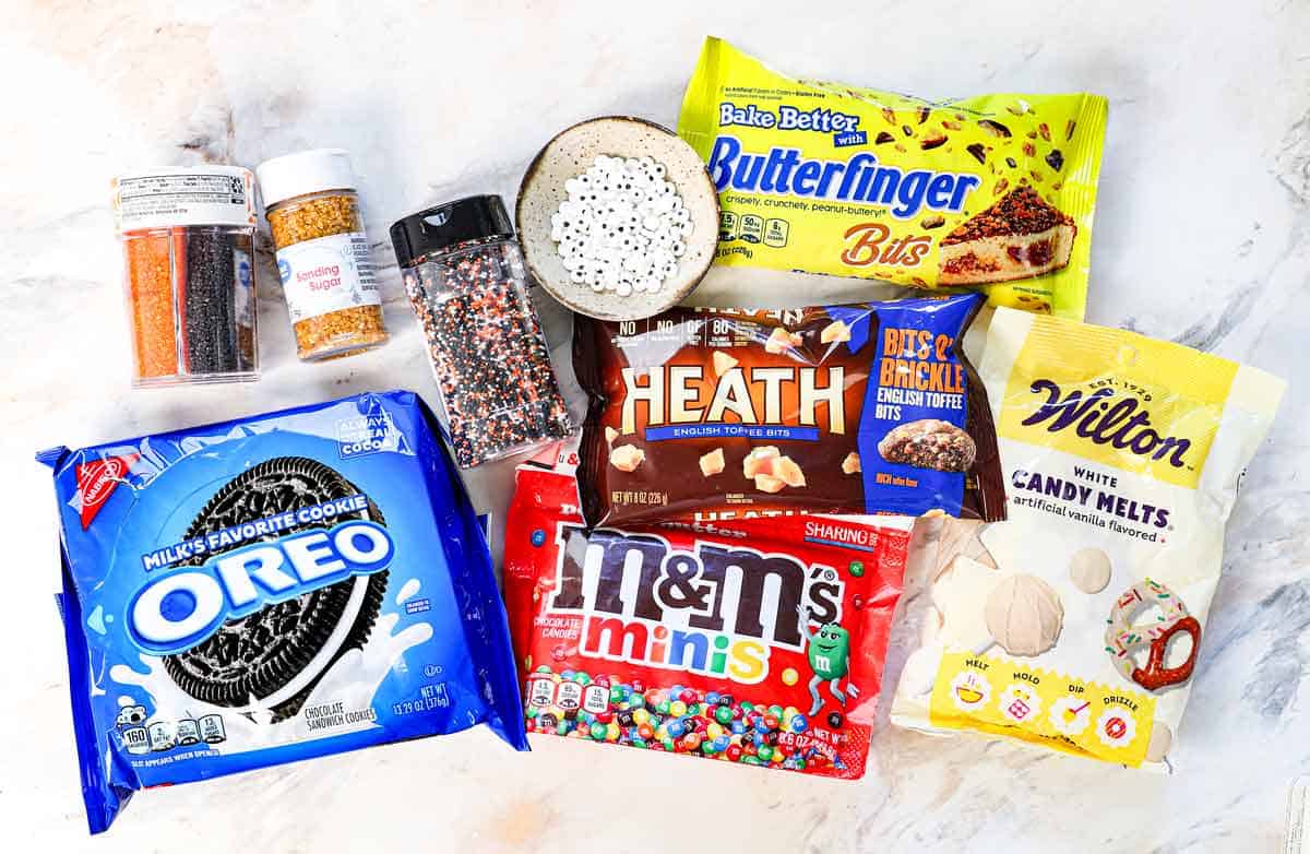 Top view of caramel Apple Toppings to decorate caramel apples with: crushed Oreos, mini M&Ms, three kinds of sprinkles, toffee bits, candy melts, candy eye balls, crushed candy