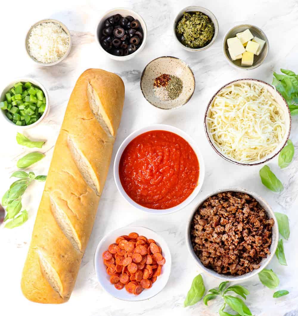 Top view of French Bread Pizza ingredients: French bread, pizza sauce, mozzarella cheese, Parmesan cheese, spices, butter, pepperoni, sausage, bell peppers and olives